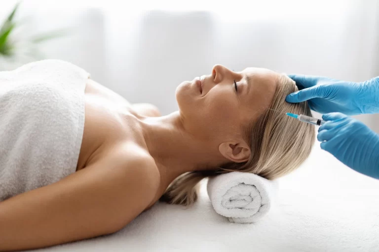 A woman lying on a spa bed with a towel around her body receives a cosmetic injection in her forehead from a professional wearing blue gloves.