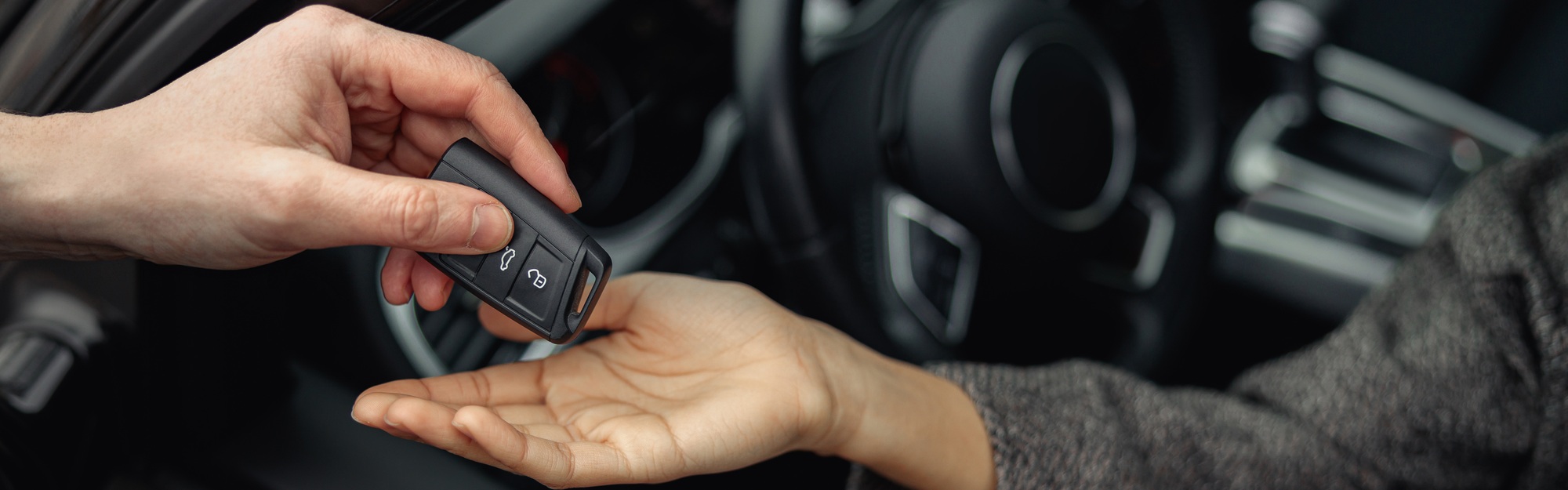 A close-up of one person handing a car key fob to another person inside a vehicle, with a steering wheel and dashboard visible in the background.