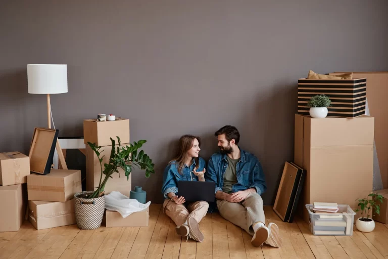 A couple sits on the floor surrounded by moving boxes, a lamp, plants, and home decor. They smile at each other while using a laptop, appearing relaxed and happy in their new home.
