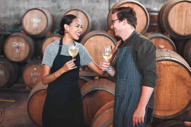 Two people wearing aprons stand in front of large wine barrels, smiling and holding glasses of white wine, appearing to enjoy a conversation in a winery setting.