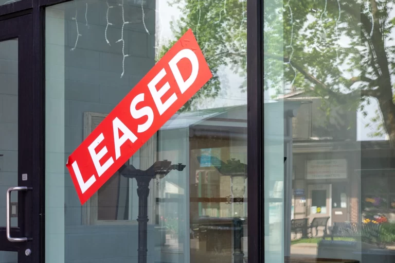 A large red and white “LEASED” sign is displayed diagonally across the glass window of a storefront. Reflections of trees and nearby buildings are visible in the window.