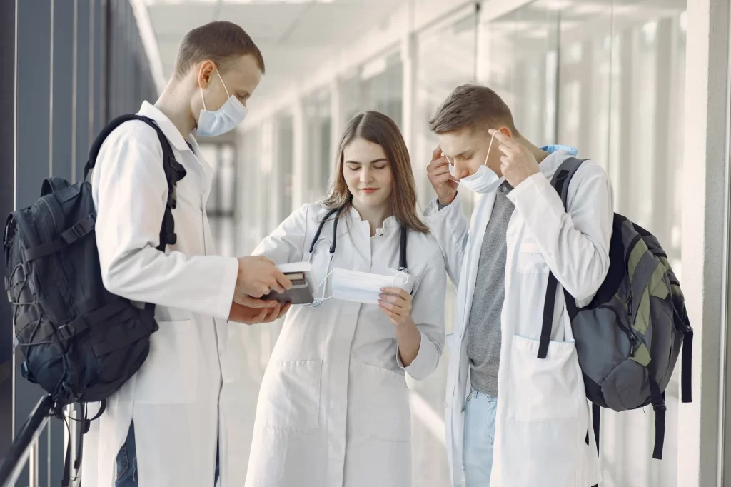 Three young medical professionals in white coats with backpacks stand in a hallway. One holds a tablet, another holds a face mask, and the third is putting on a mask. All appear to be discussing something.