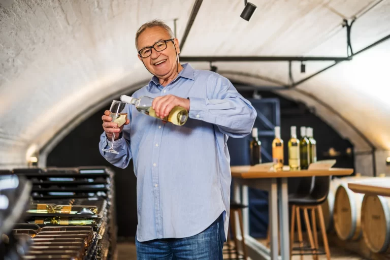 An older man in a light blue shirt smiles while pouring white wine into a glass in a wine cellar, surrounded by bottles and wine barrels.